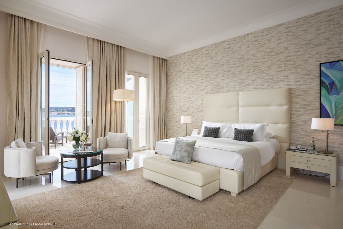 A modern hotel room with a large bed, a seating area with two chairs, a small round table, and a view through the open curtains.