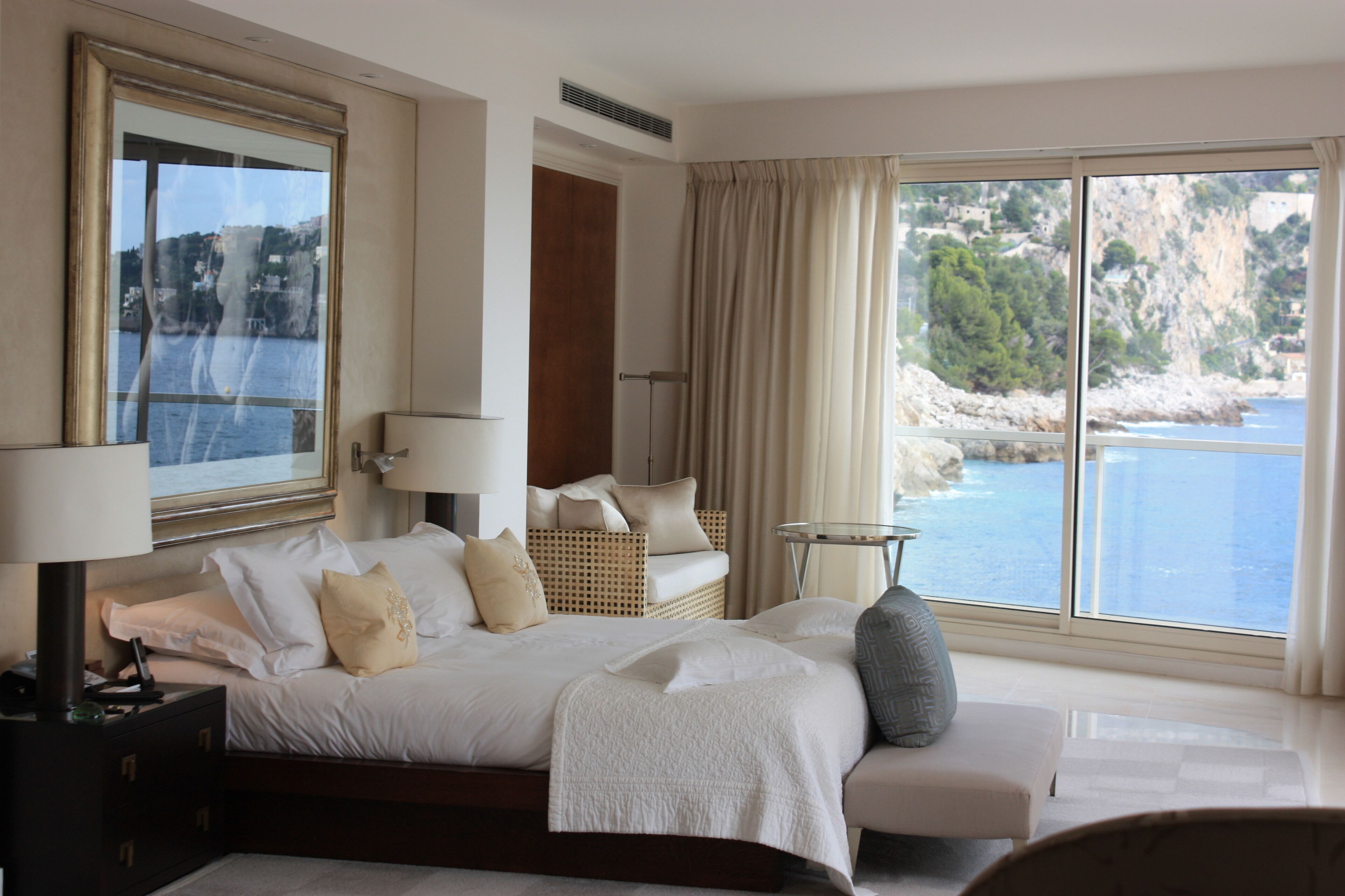 A spacious bedroom with a large bed, a bedside table, a chair, and a view of the sea through large windows.