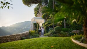 A couple walking on a stone pathway surrounded by lush greenery and a stone wall. - Hôtel Cap-Estel (Èze)