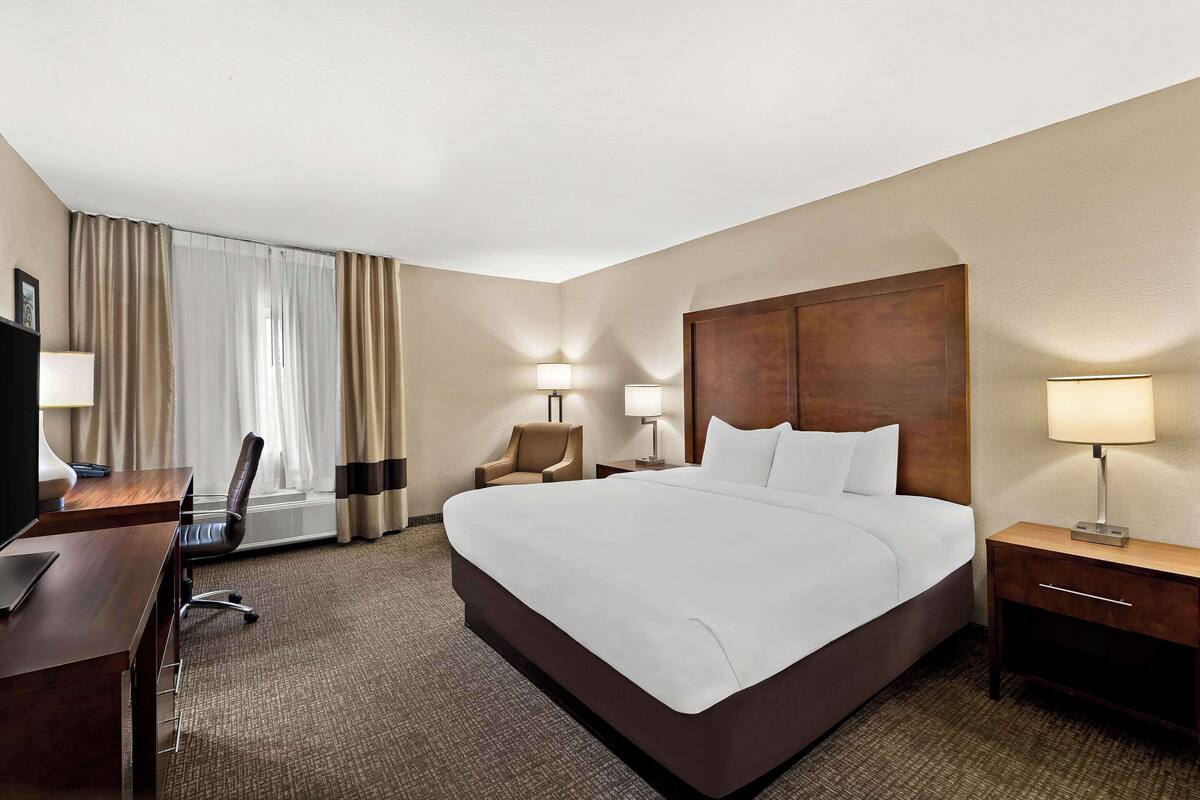A hotel room with a large bed, a desk with a chair, a nightstand with a lamp, and a window with curtains.