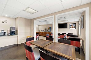 A hotel dining area with multiple tables and chairs, a kitchenette, and a television mounted on the wall.