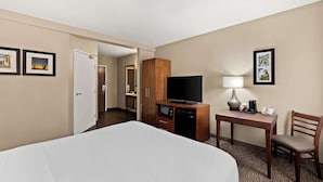 Habitación de hotel con cama, escritorio con lámpara, televisor y una silla. En la pared, hay cuadros enmarcados y una puerta que da a otra habitación.