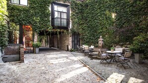 A courtyard with a cobblestone path, outdoor seating area with metal chairs and tables, and a brick building partially covered with ivy. - Hotel de Castillion (Bruges)