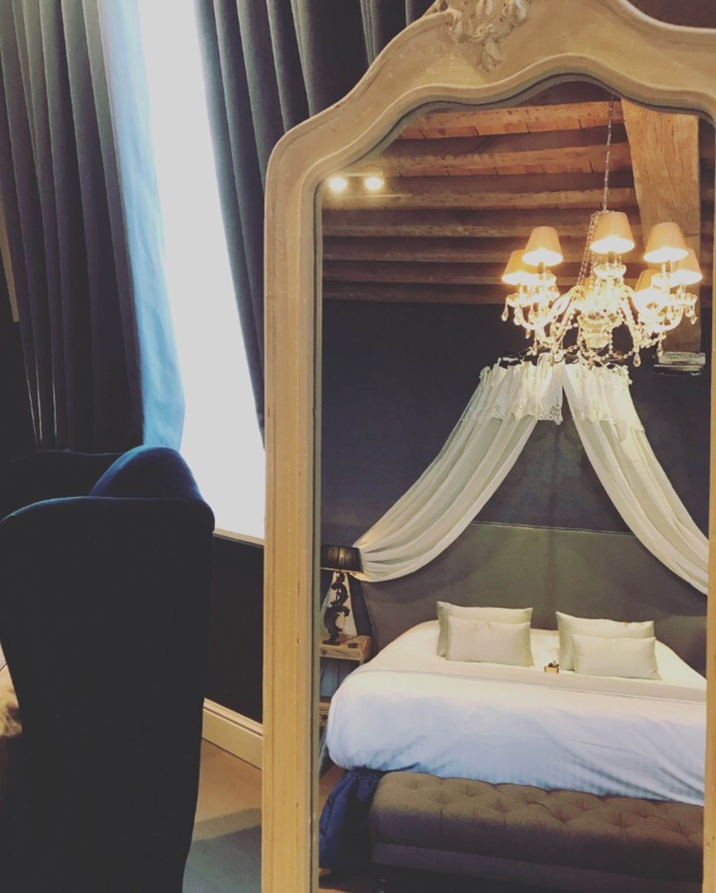 Un dormitorio con un espejo grande, una cama con ropa de cama blanca, una araña y un cabecero de color oscuro.