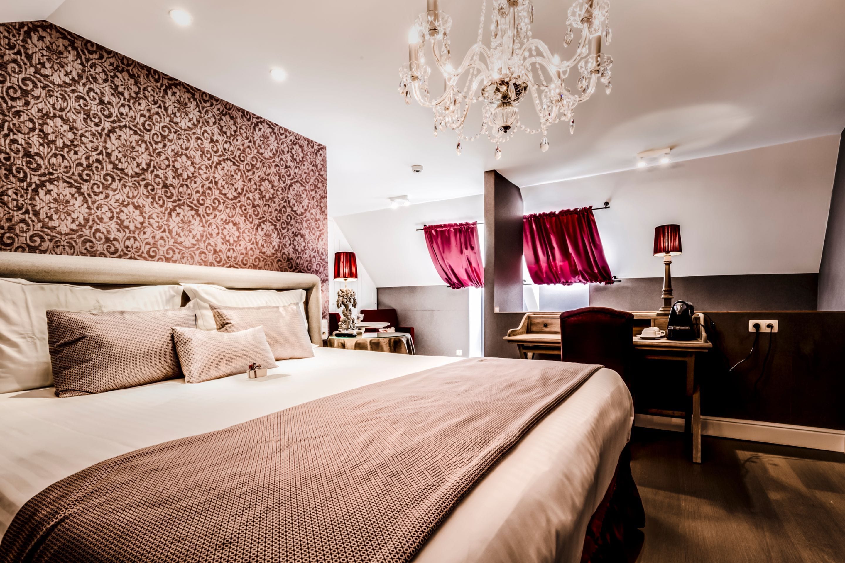 A hotel room with a large bed, a desk, a chair, a chandelier, and a patterned wall.
