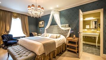 Een slaapkamer met een groot bed, een baldakijn, een kroonluchter, een nachtkastje, een stoel en een badkamer met een wastafel en spiegel.
