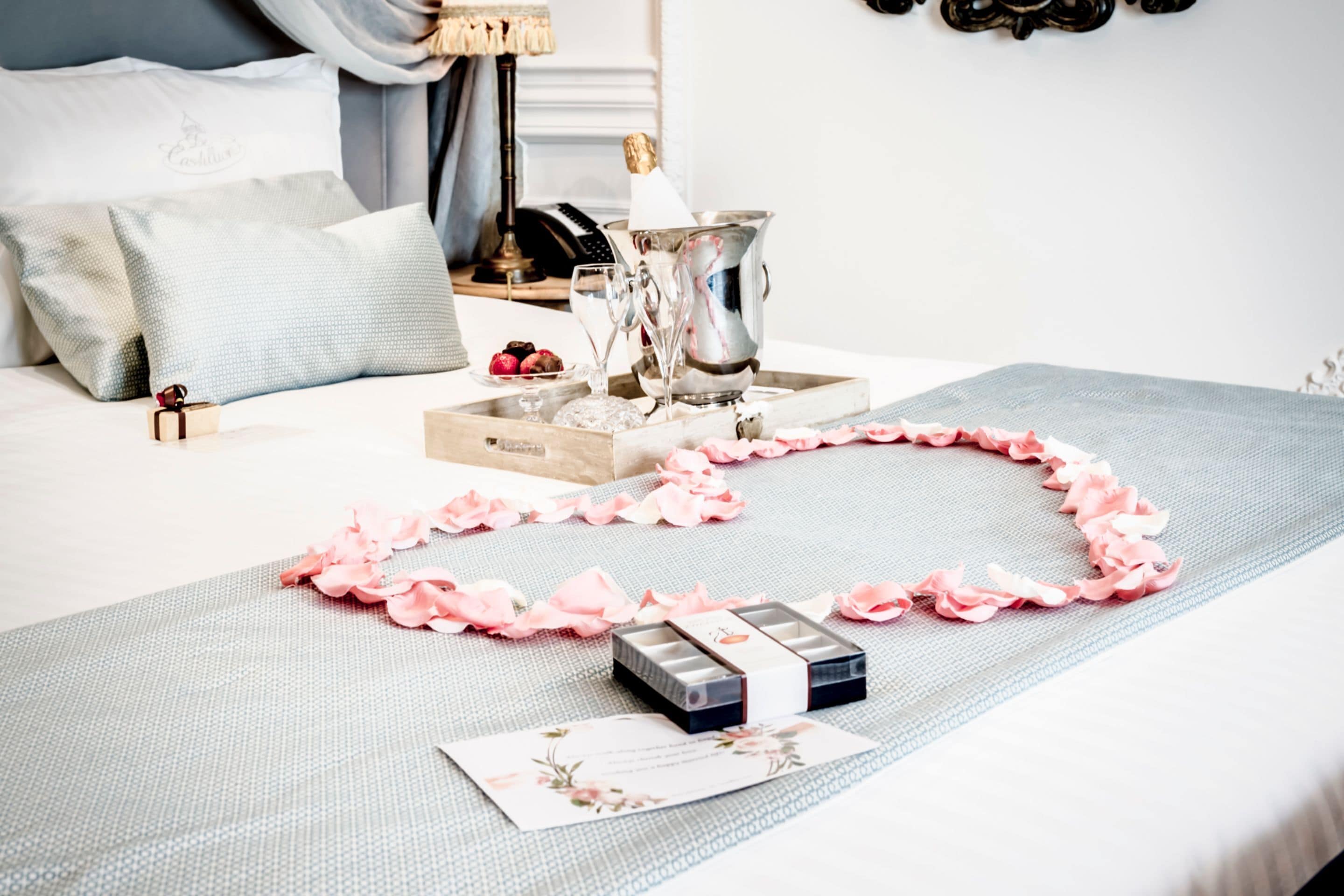 A neatly arranged bedroom with a bed, pillows, a bedside table with a lamp, a champagne bottle, glasses, and a tray with chocolates and flowers.