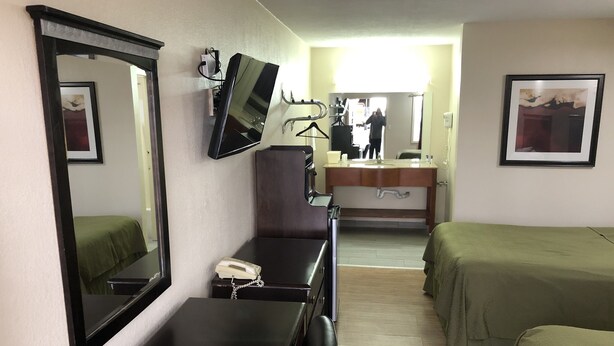 A hotel room with a bed, a desk with a telephone, a mirror, a TV, and a bathroom visible in the background.