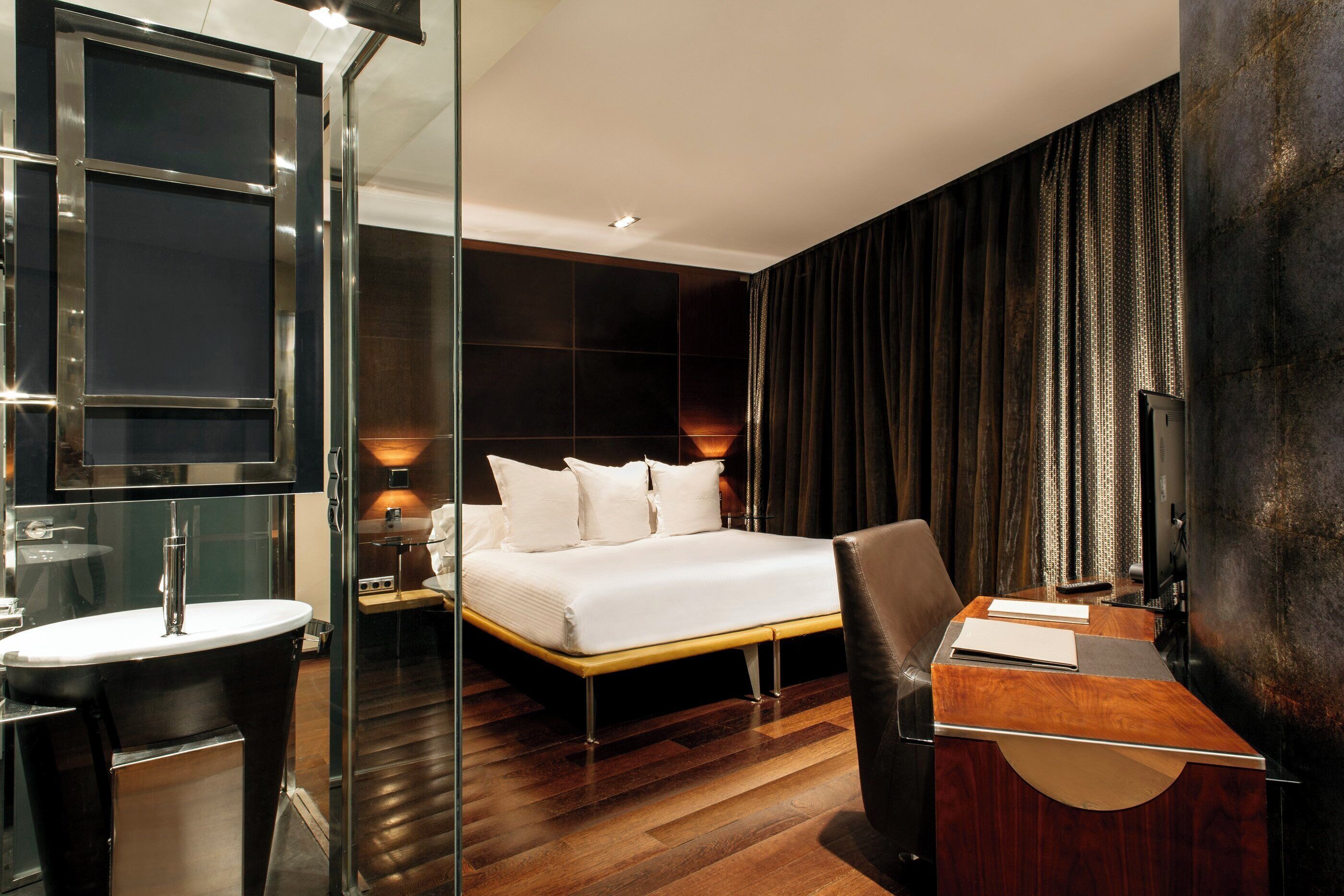 A modern hotel room with a large bed, a bedside table, a desk, and a mirror.