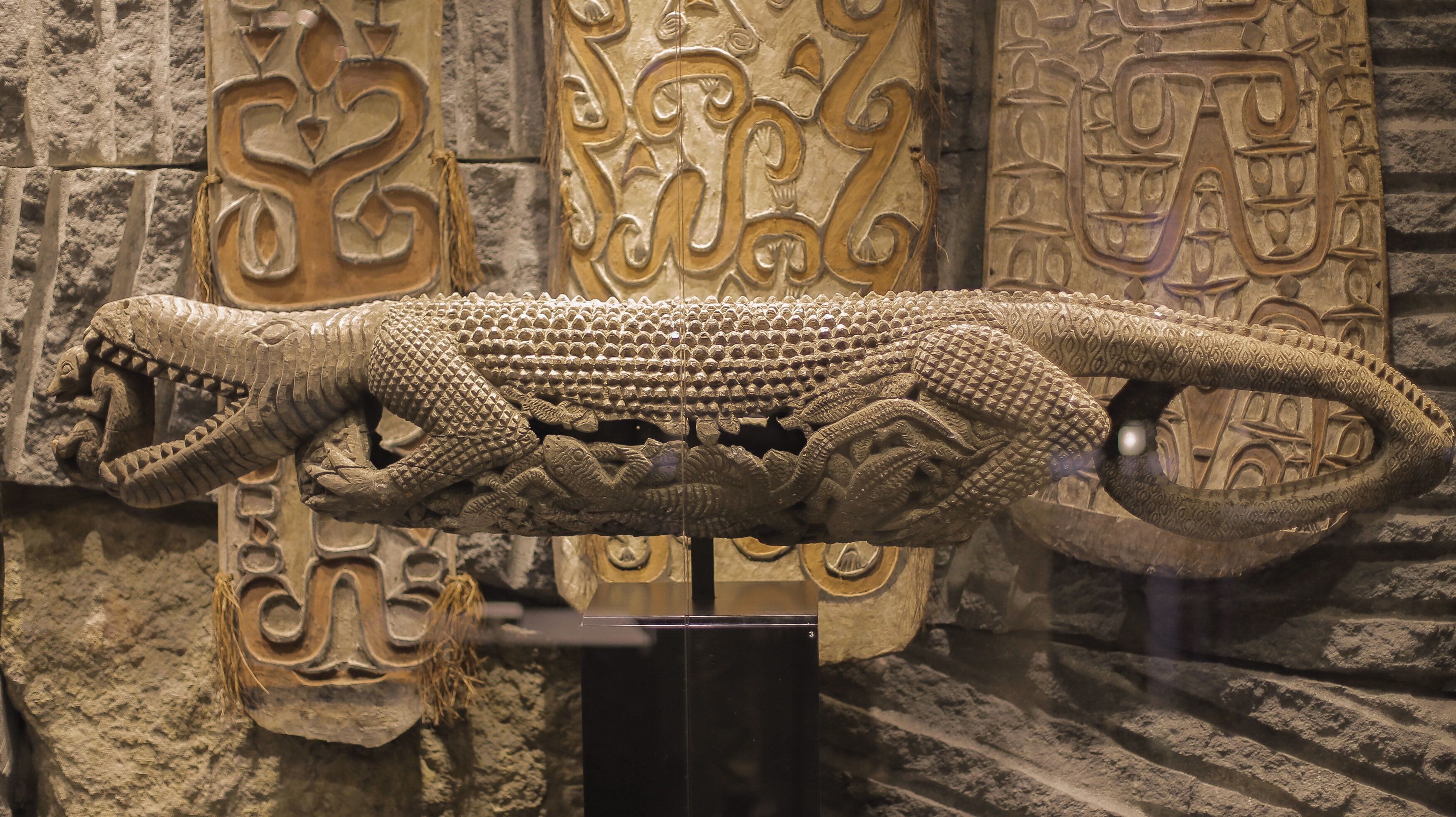 A large, intricately carved stone sculpture of a reptile, possibly a crocodile, with detailed scales and claws, displayed against a backdrop of similarly ornate stone carvings.