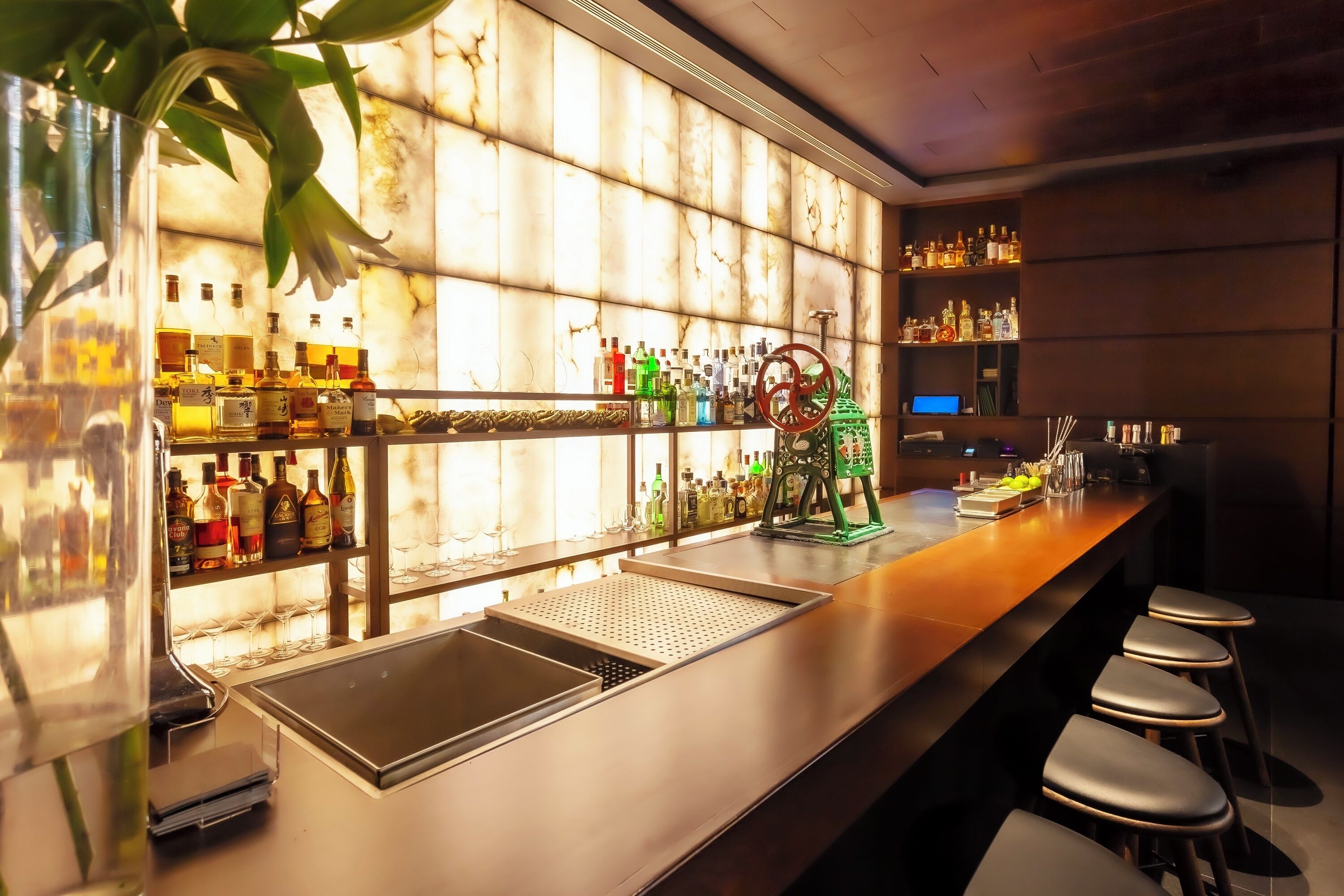Uma área de bar com prateleiras cheias de garrafas de bebidas, um balcão, banquetas e uma planta decorativa.