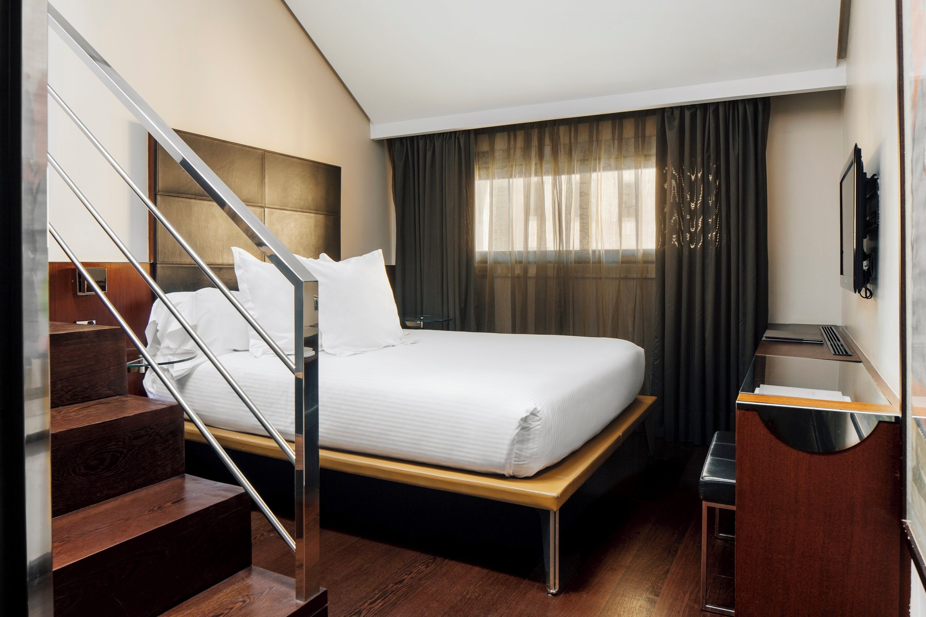 Una habitación de hotel con cama, escritorio, silla, escalera y ventana con cortinas.