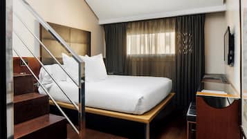 Una habitación de hotel con cama, escritorio, silla, escalera y ventana con cortinas.