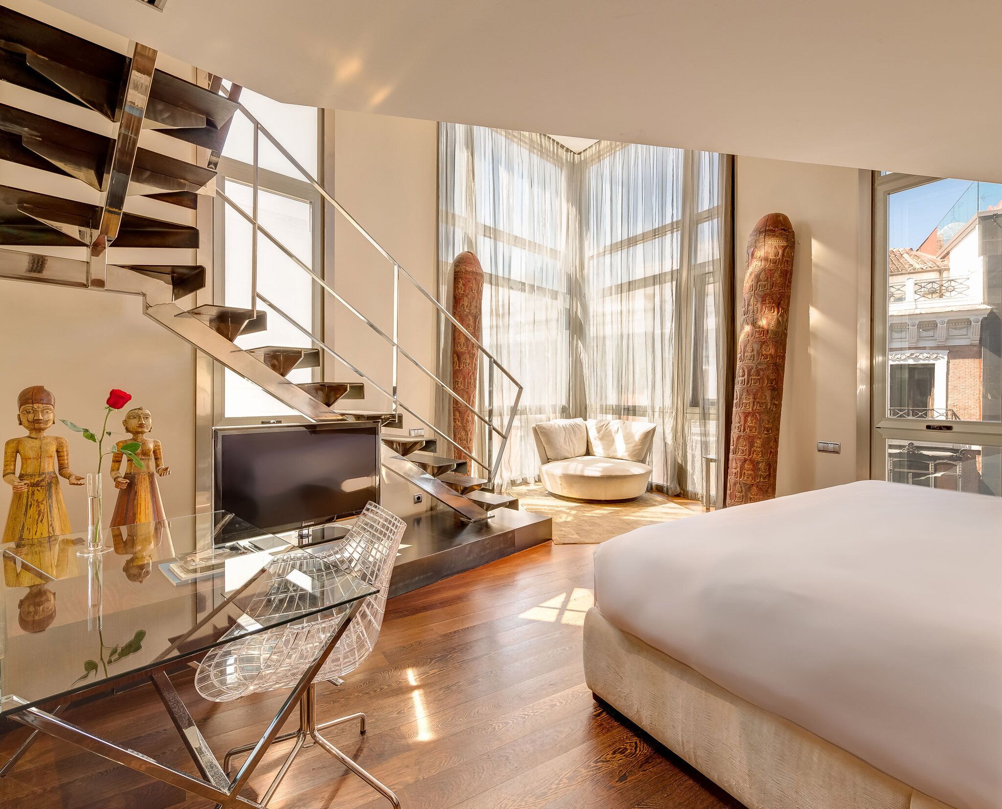 Una habitación de hotel moderna con un escritorio de vidrio, una cama, un sofá y una escalera con barandillas de metal.