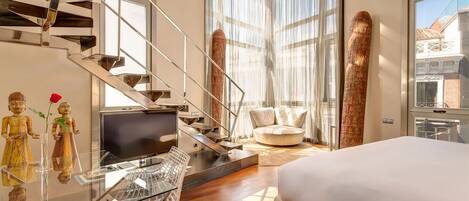 Una habitación de hotel moderna con un escritorio de vidrio, una cama, un sofá y una escalera con barandillas de metal.