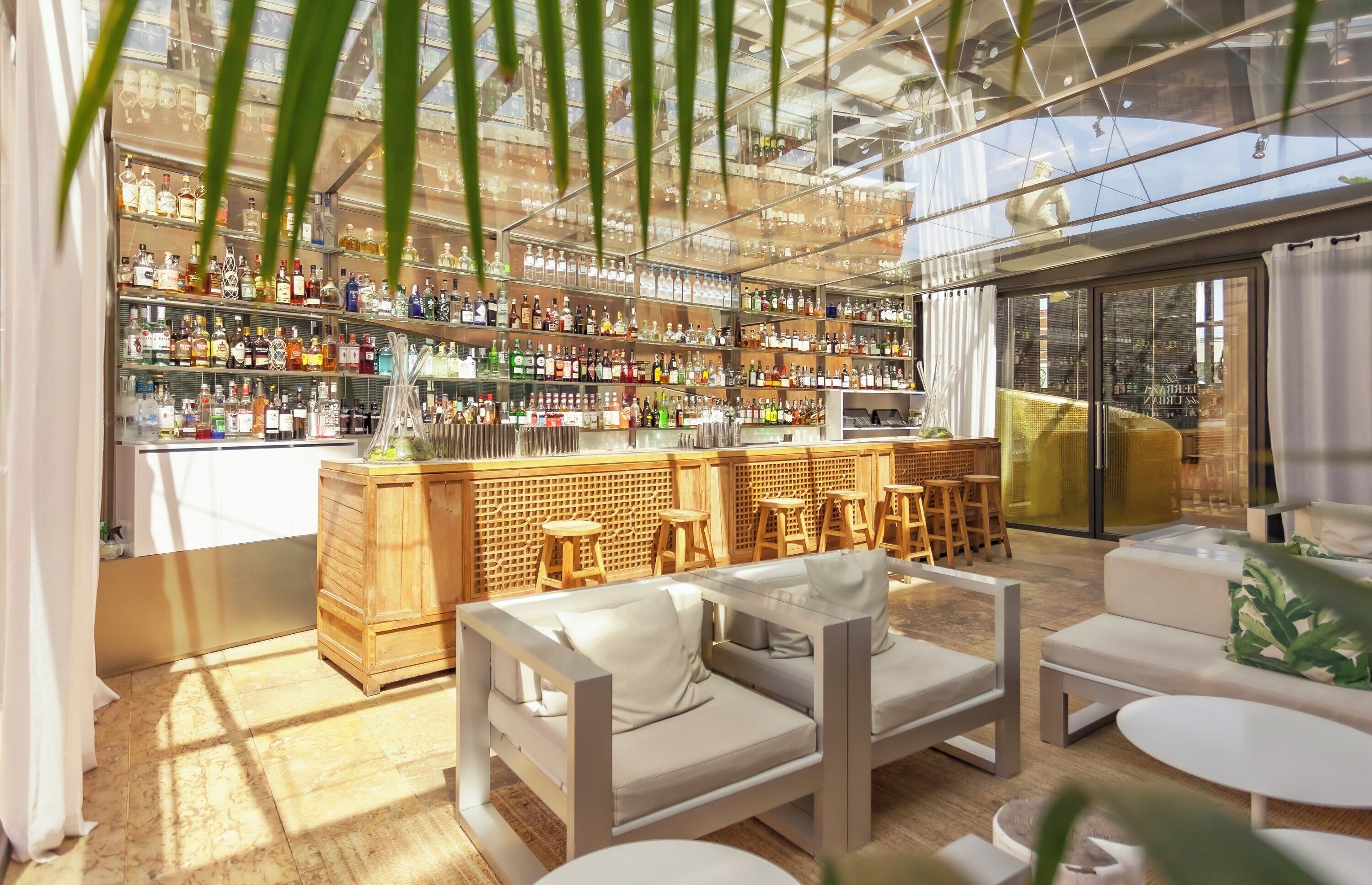 Zona de bar moderna con una variedad de botellas de licor, mobiliario de madera y techo de cristal.