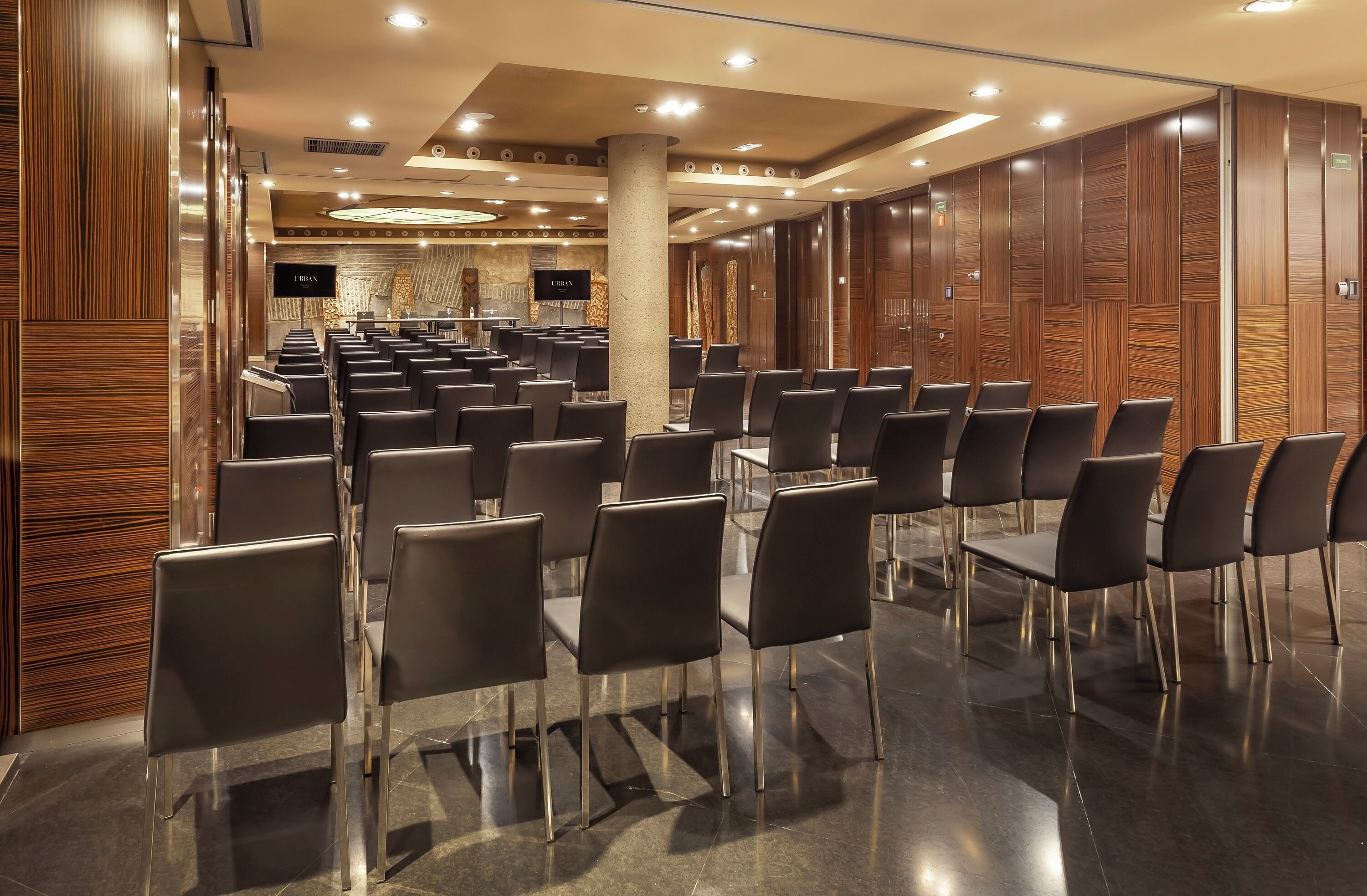 Un salón de conferencias con hileras de sillas negras, paneles de madera en las paredes y una pantalla grande al frente.