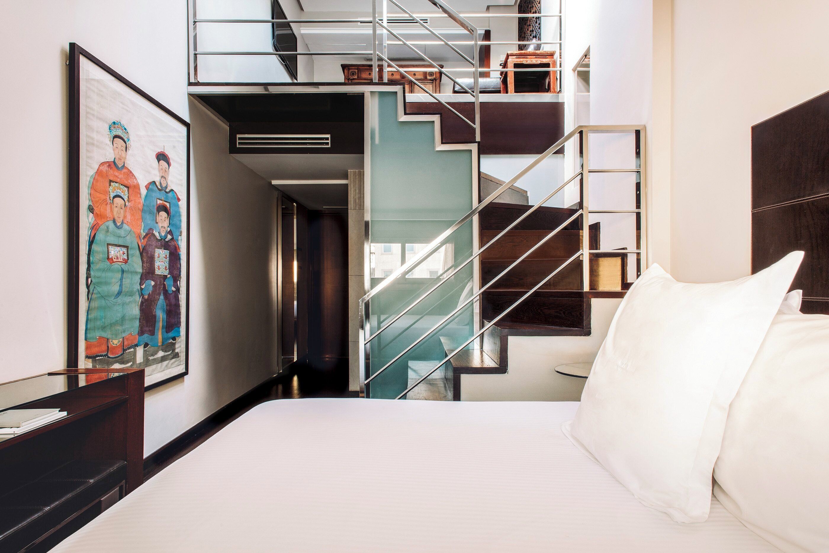 Una habitación de hotel con una cama, un cuadro, una escalera y vistas al pasillo.
