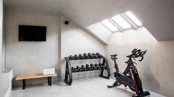 Een sportschool met een plafondventilator, een aan de muur bevestigde televisie, een bank, een dumbbellrek en een hometrainer.