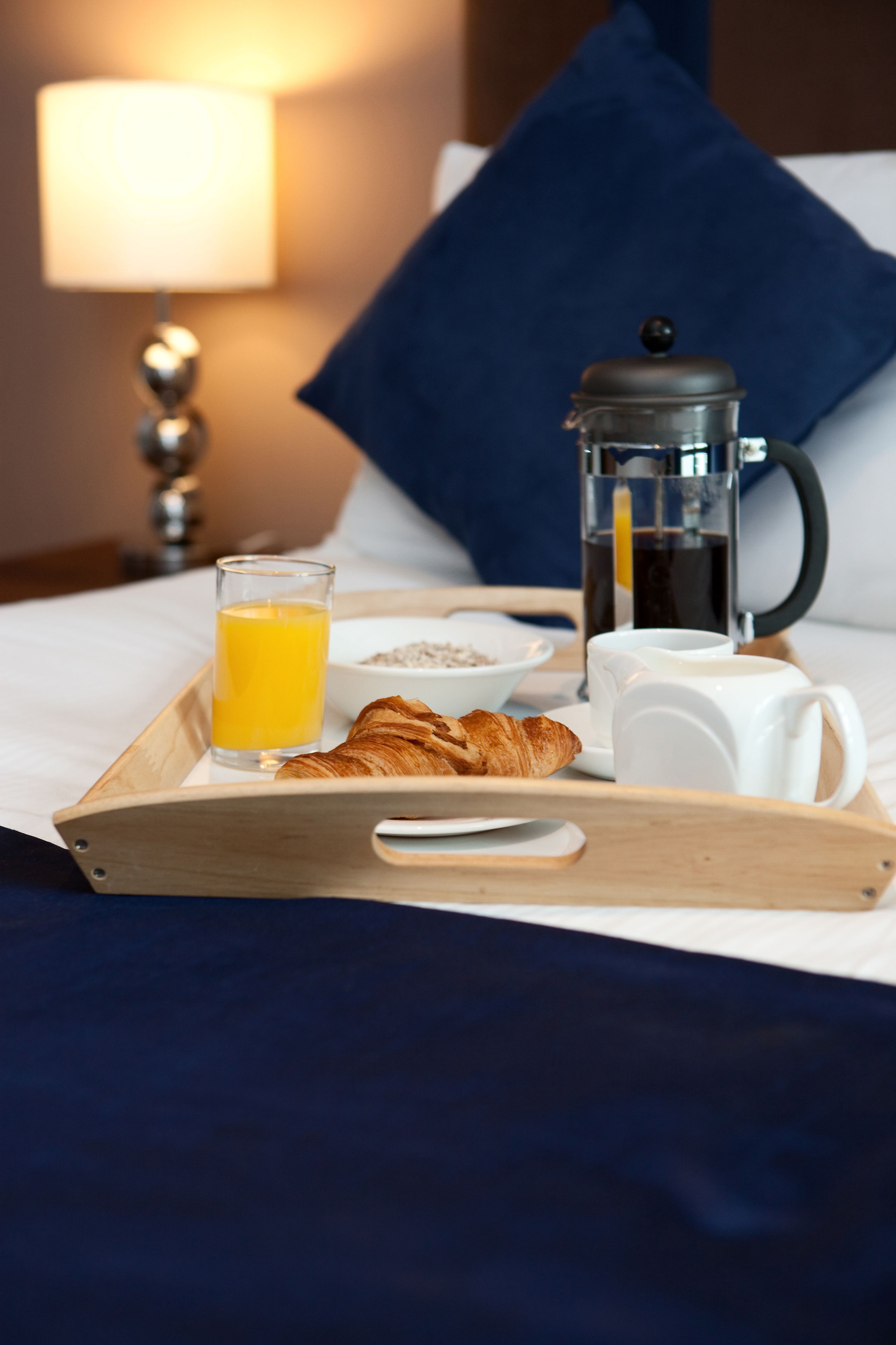 Bandeja de café da manhã com croissants, um copo de suco de laranja, uma cafeteira e uma xícara sobre a cama, com um travesseiro azul e uma luminária ao fundo.