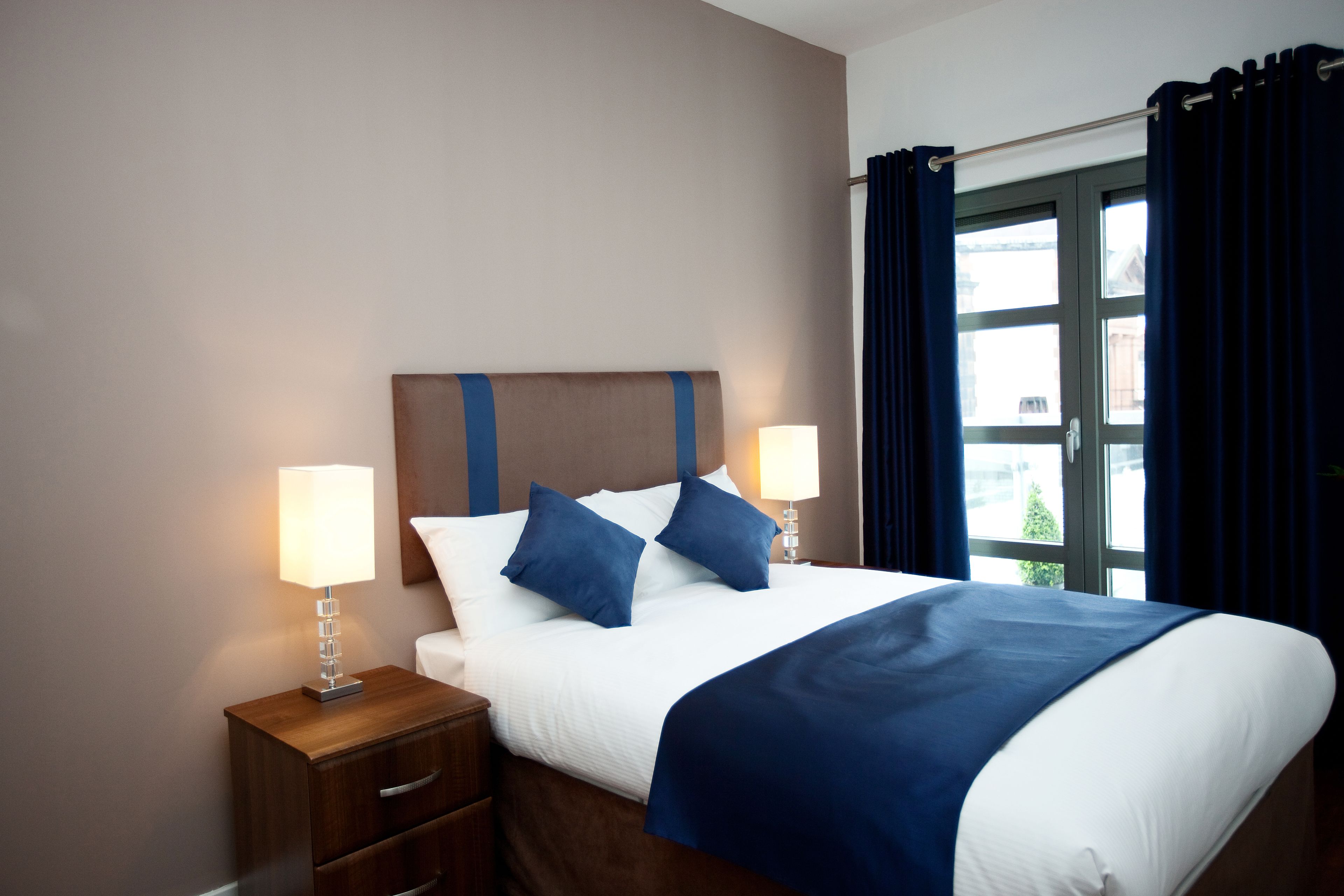 A neatly made bed with a blue headboard, two bedside lamps, and a window with dark curtains.