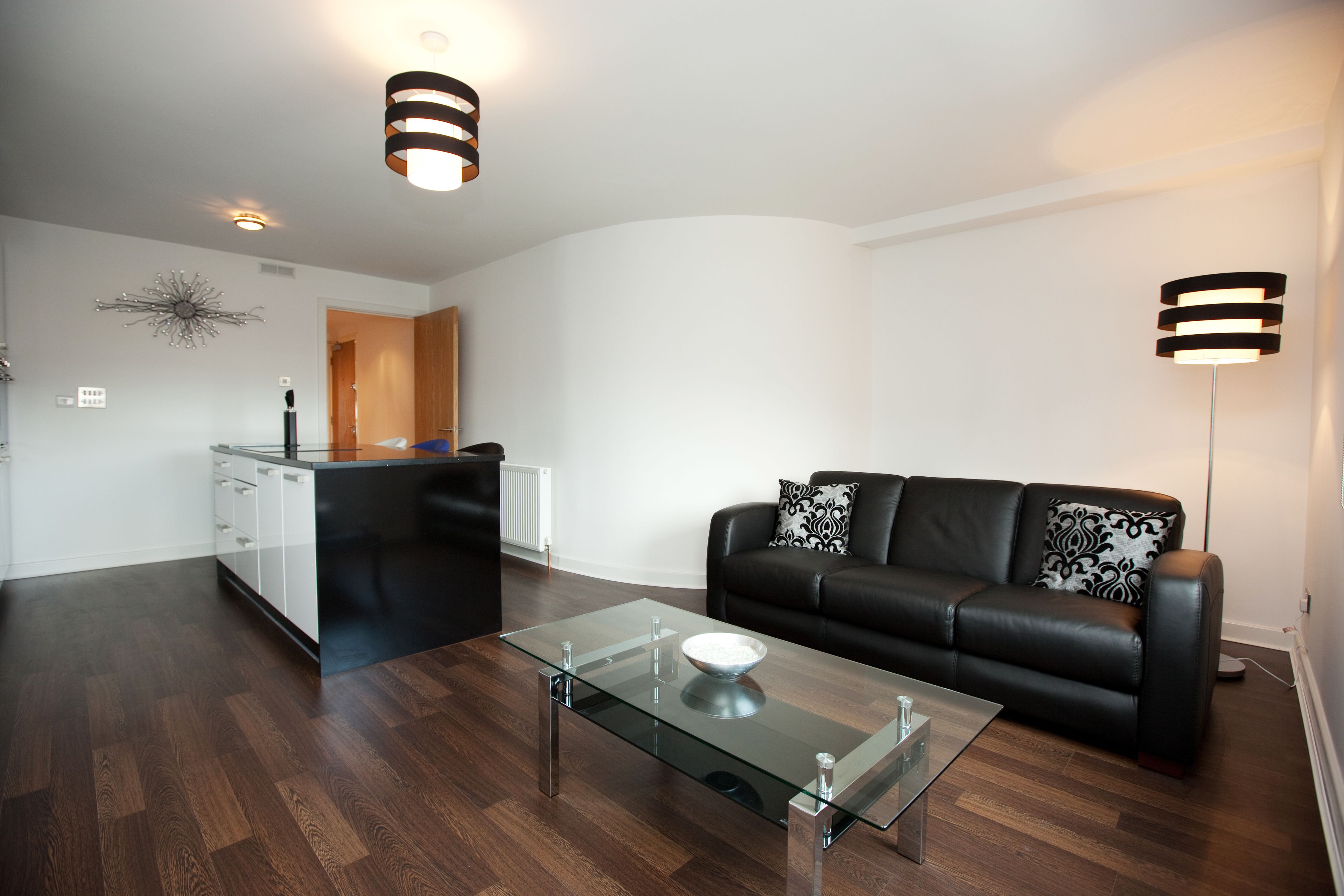 Una sala de estar moderna con una mesa de centro de vidrio, un sofá de cuero negro con cojines estampados, y una zona de cocina al fondo.