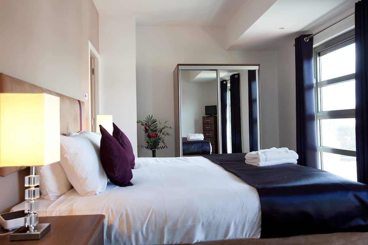 A hotel room with a bed, bedside table, lamp, and a view of the city through large windows.