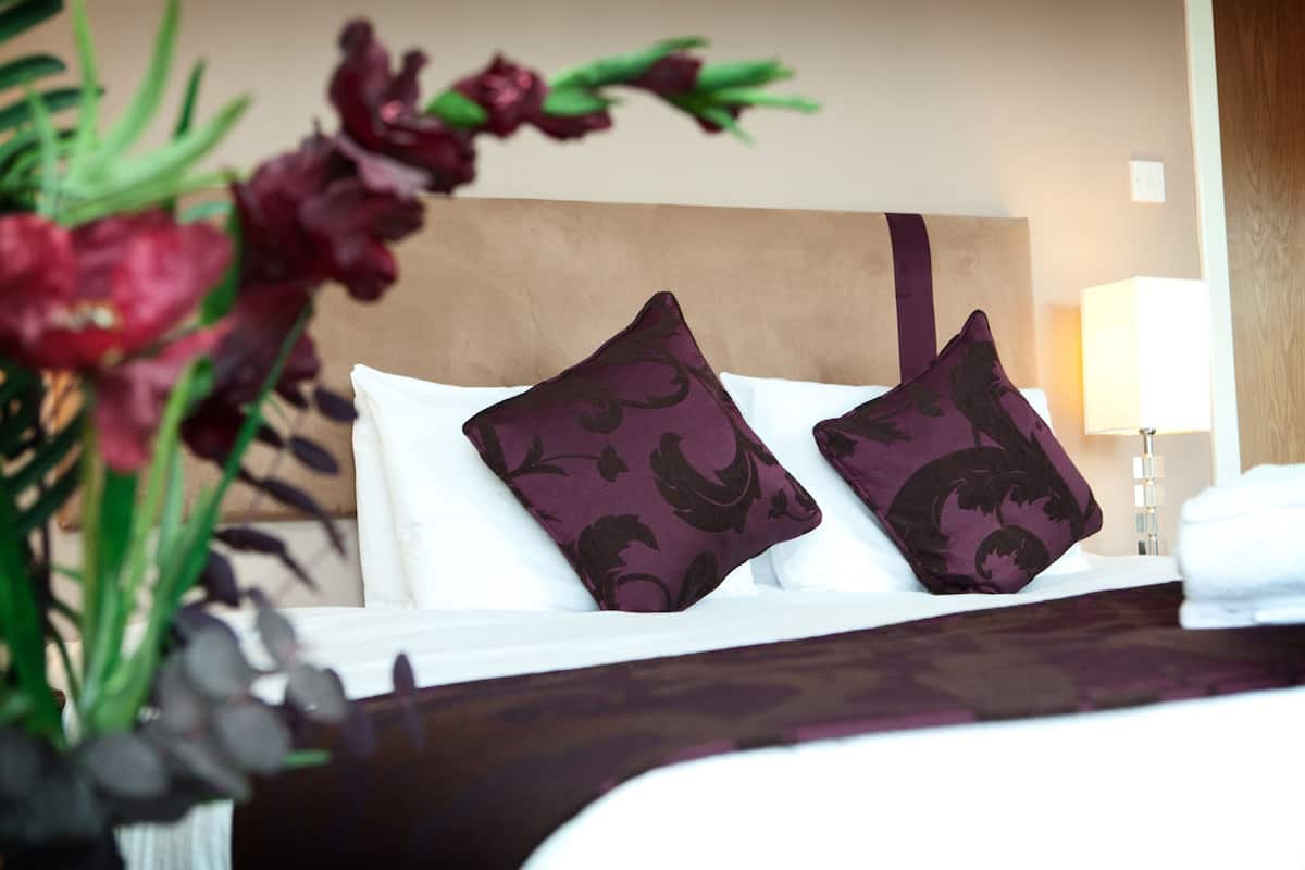 A hotel room with a bed, two decorative pillows, a bedside lamp, and a vase with flowers.