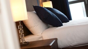 A neatly made bed with white linens and dark blue pillows, a bedside table with a lamp, and a window with curtains in the background. - The Spires Serviced Apartments Glasgow (Glasgow)