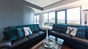 A modern living room with two black leather sofas, each with plaid cushions and a deer pillow, a glass coffee table with remote controls, and large windows offering a city view. - The Spires Serviced Apartments Glasgow (Glasgow)
