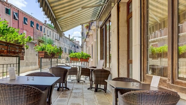 An outdoor café with wicker tables and chairs, potted plants, and a tiled floor. - Hotel Hesperia (Venice)