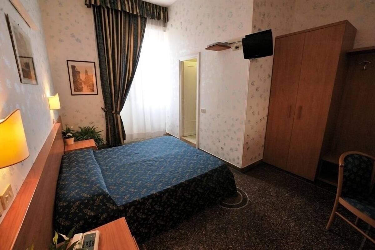 A hotel room with a bed, a desk, a chair, a wardrobe, a lamp, and a picture on the wall.