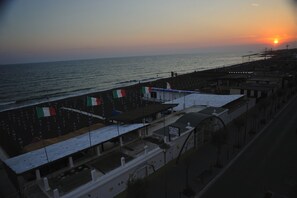 A coastal view with a sunset, a pier, and buildings with flags.