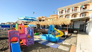 Un parque infantil en la playa con estructuras de juego coloridas y un edificio con balcones y banderas al fondo.