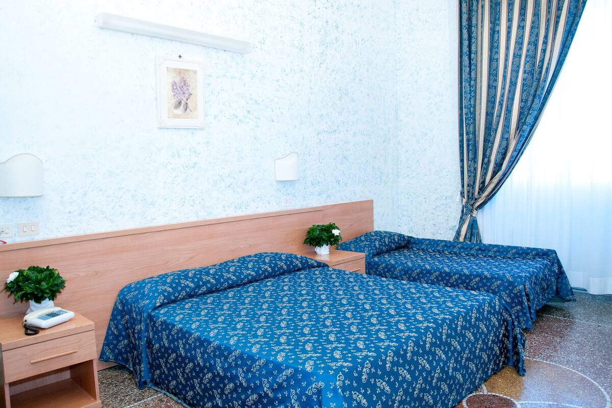 A hotel room with two beds, a bedside table with a plant, a wall-mounted light, and a window with curtains.