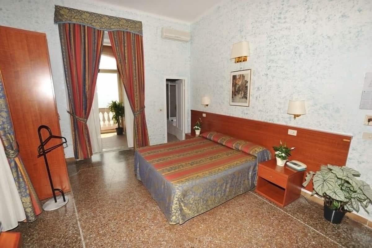 A hotel room with a bed, bedside tables, a nightstand, a potted plant, and a door leading to another room.
