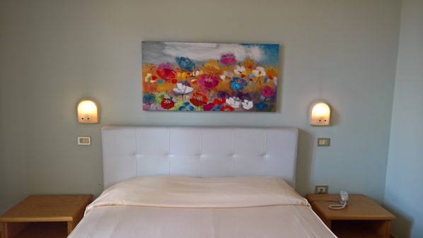 Een slaapkamer met een wit hoofdeinde, een kleurrijk bloemen schilderij aan de muur en twee bedlampen.