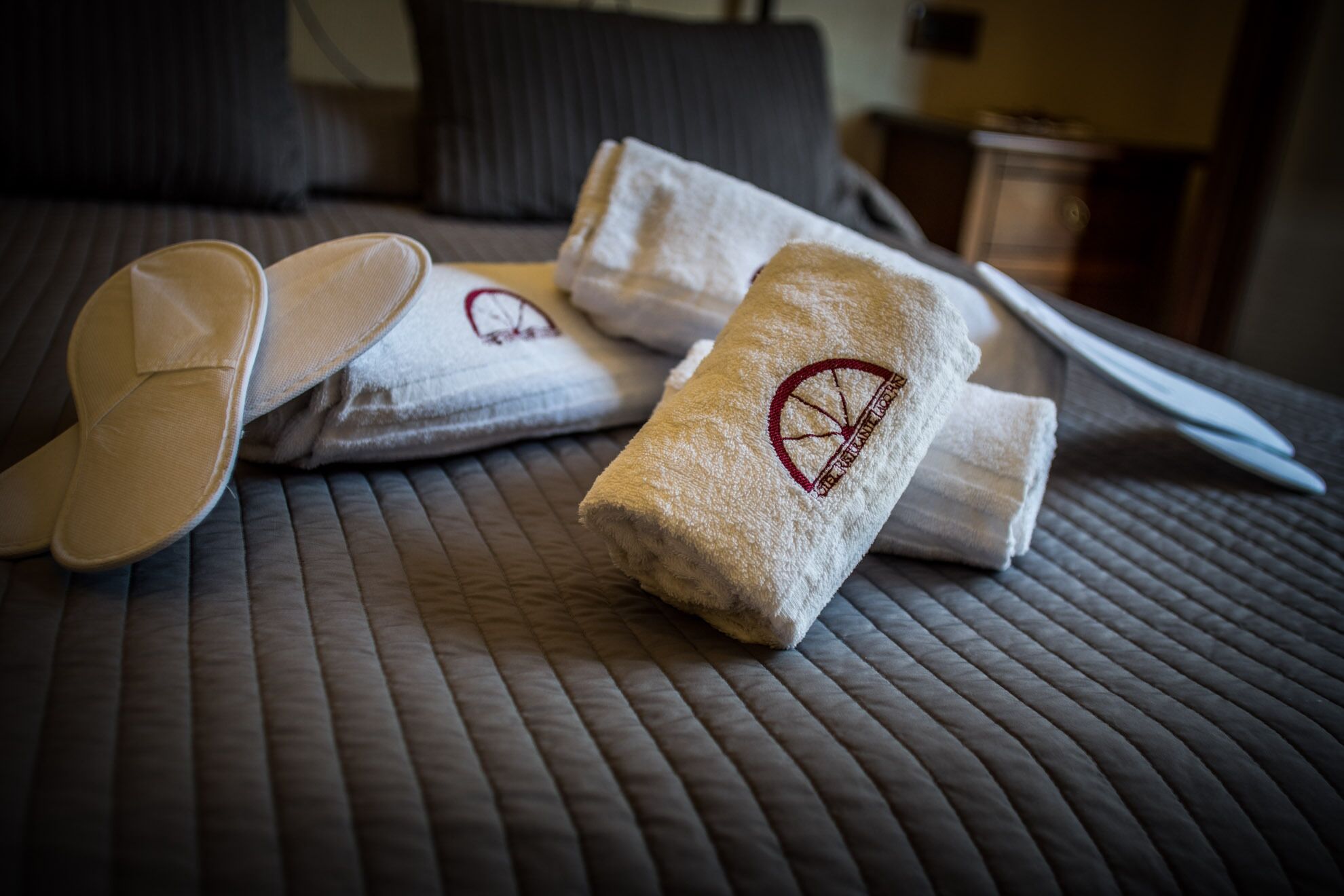 A neatly made bed with a quilted cover, two folded towels with a logo, and a pair of slippers.
