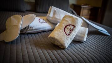 A neatly made bed with a quilted cover, two folded towels with a logo, and a pair of slippers.