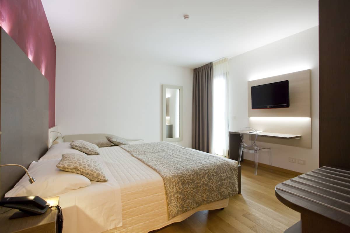 A modern hotel room with a large bed, a bedside table with a lamp, a flat-screen TV mounted on the wall, and a desk with a chair.