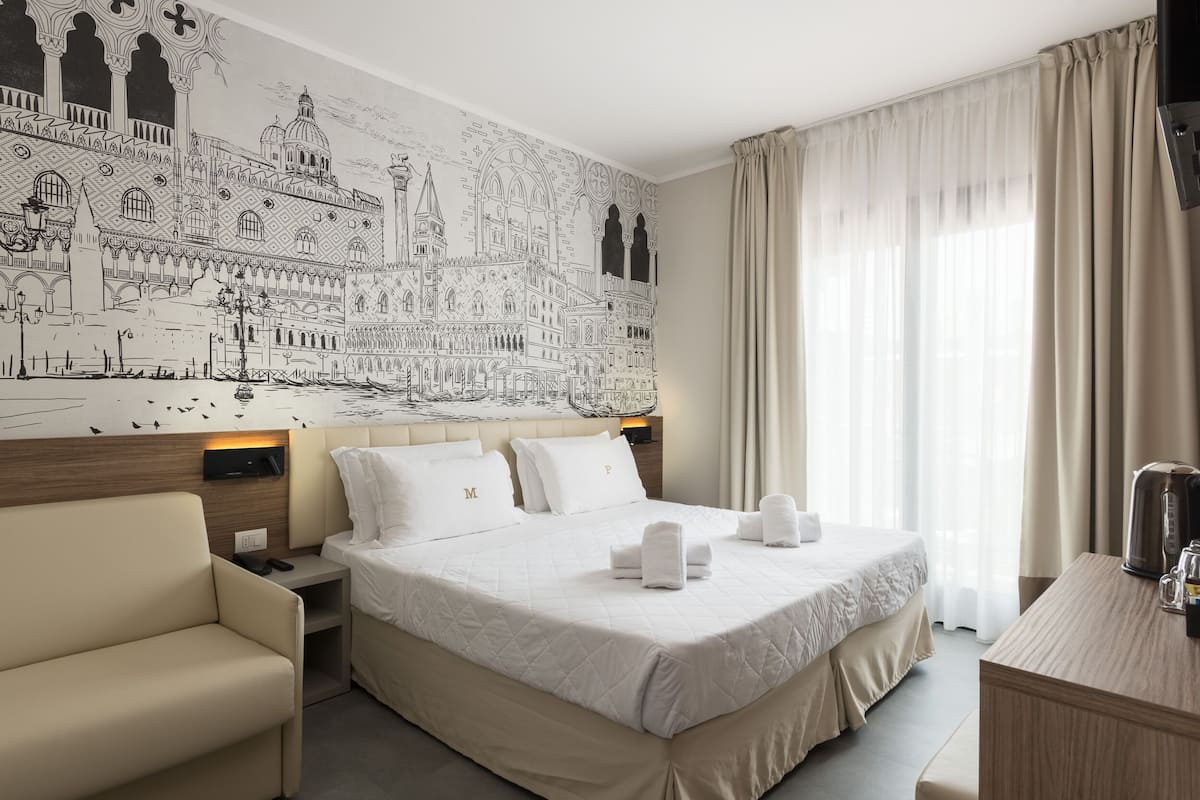 A hotel room with a large bed, a sofa, a desk, and a wall mural of a cityscape.