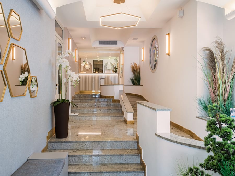 A modern hotel lobby with a marble staircase, geometric wall mirrors, and indoor plants.