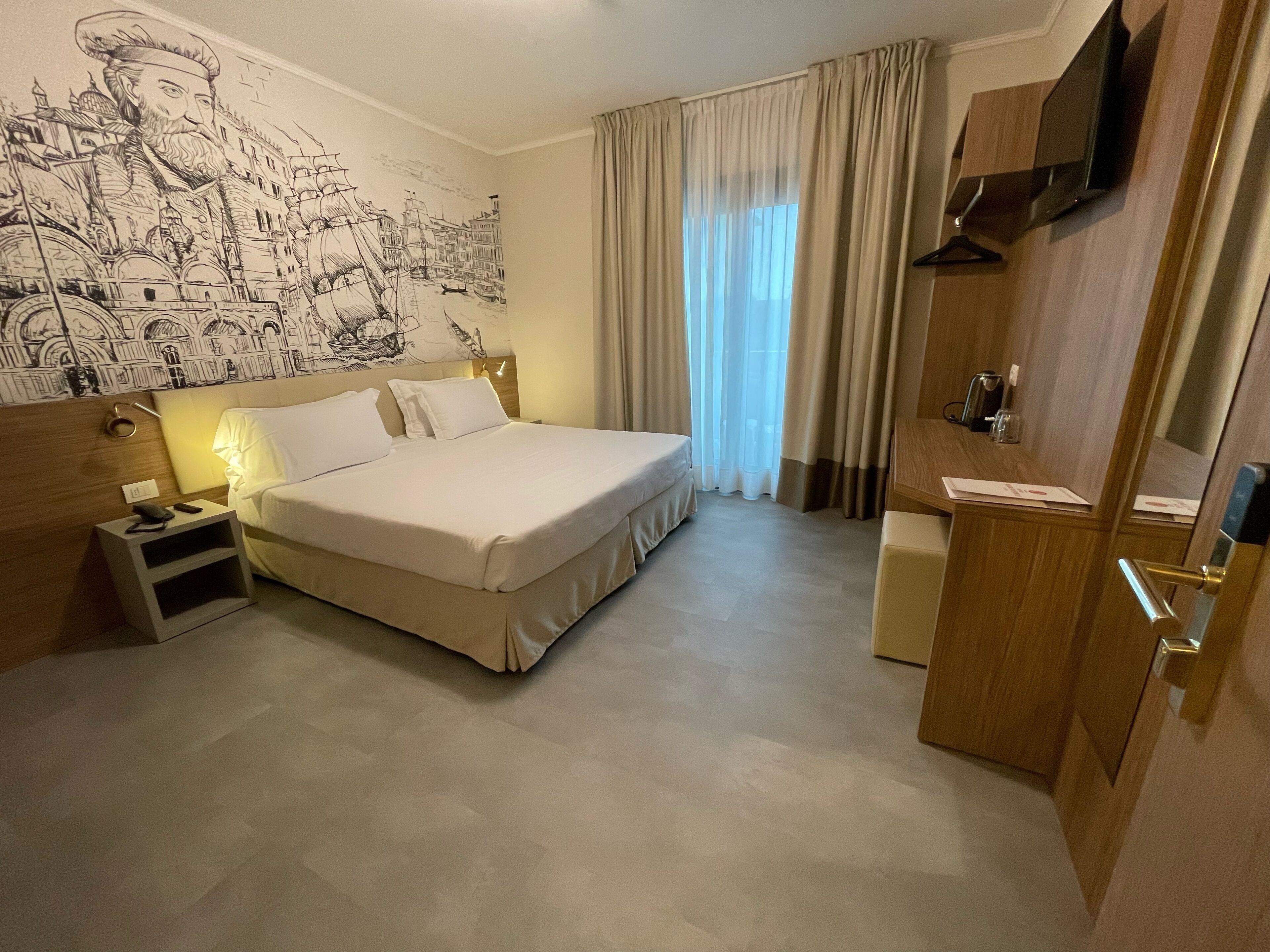 A hotel room with a large bed, a nightstand, a desk, a television, and a mural on the wall.