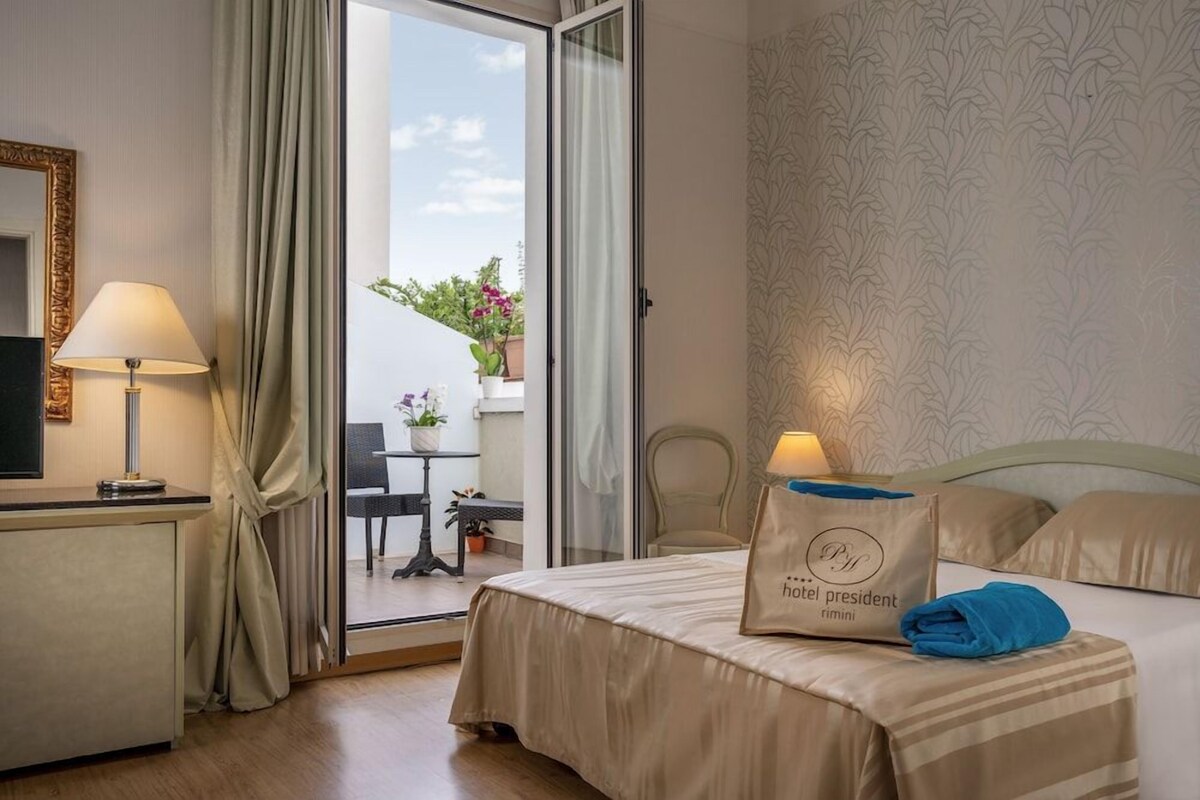 Habitación de hotel con cama, escritorio, lámpara, bolso con el nombre del presidente del hotel y vistas al exterior.