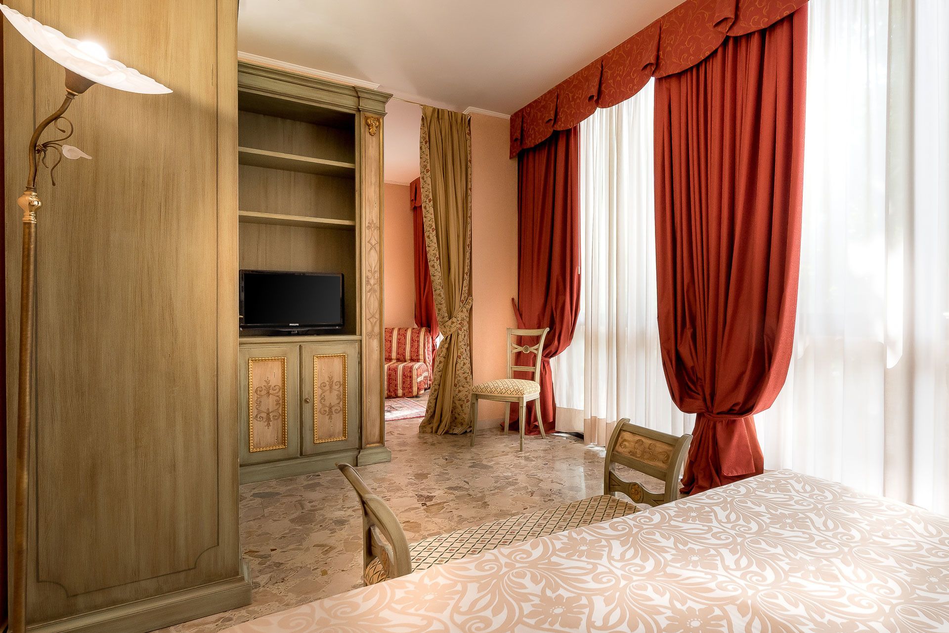 A hotel room with a television on a wooden cabinet, a bed with a patterned bedspread, a chair, and red curtains.