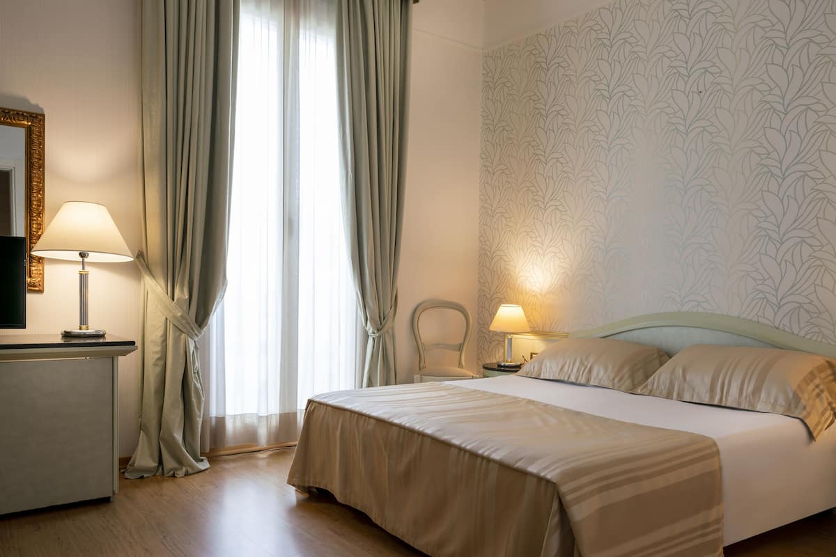 Ein Hotelzimmer mit einem Bett, zwei Nachttischlampen, einem Sessel, einem Schreibtisch mit Lampe und einem großen Fenster mit Vorhängen.