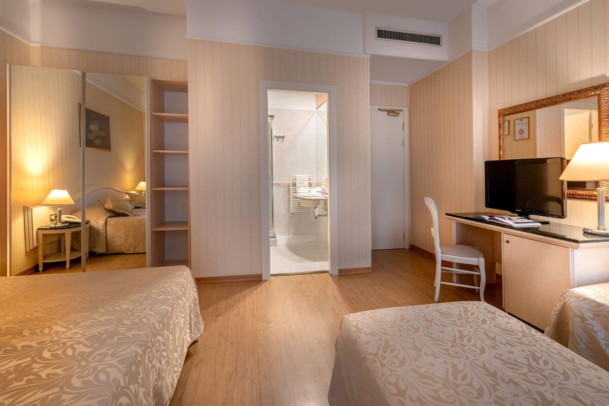 Ein Hotelzimmer mit zwei Betten, einem Schreibtisch mit Fernseher, einem Sessel, einem kleinen Tisch mit Lampe und einem Kleiderschrank.