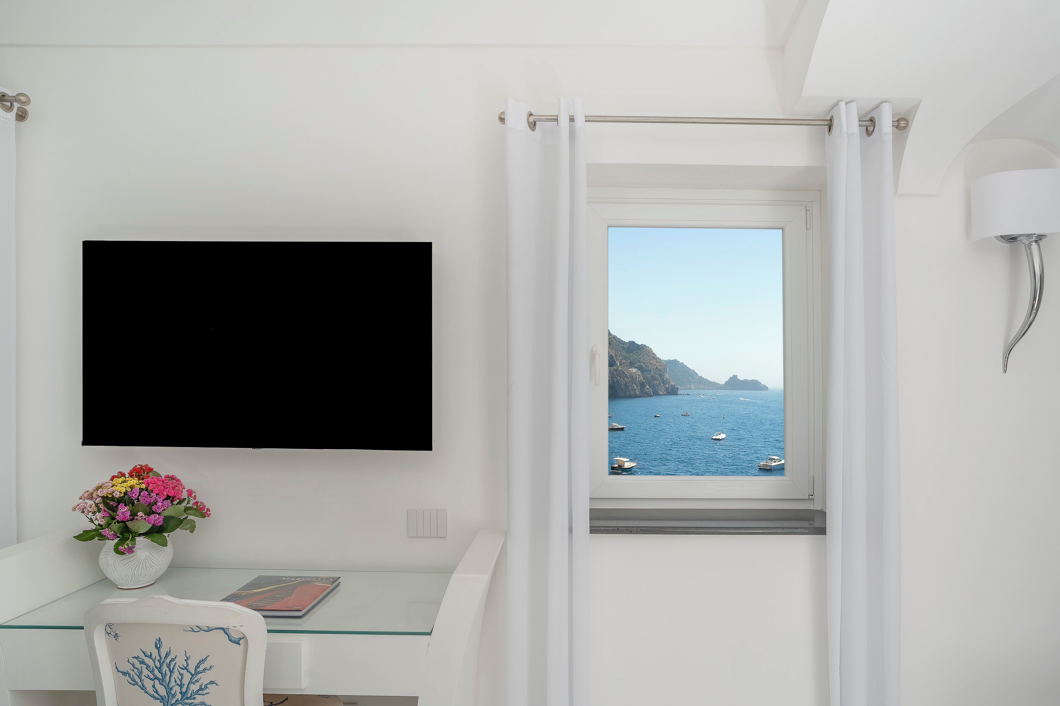 A bathroom with a glass desk, a white chair, a potted plant, a mounted TV, and a window with a view of the sea and boats.