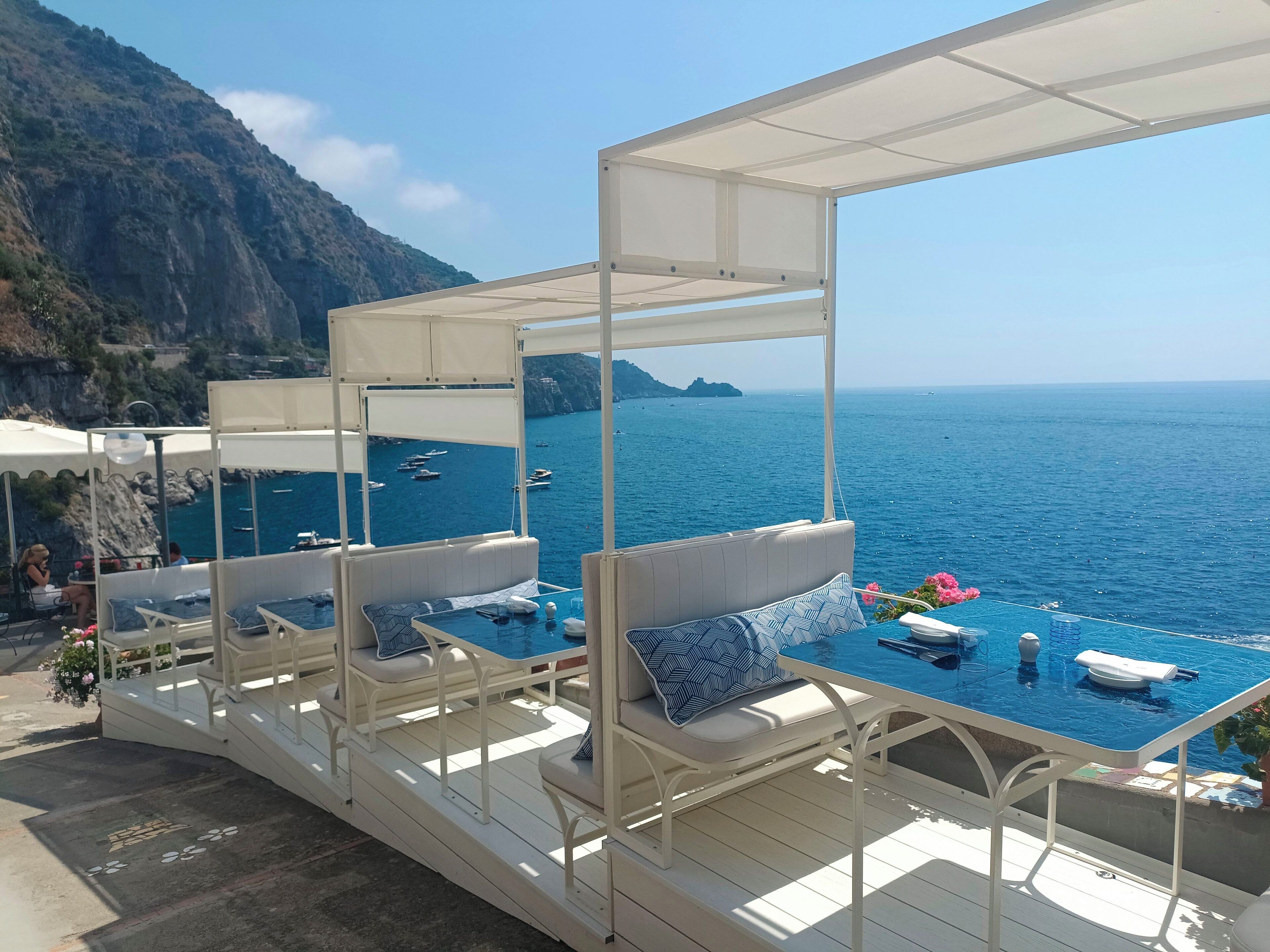 A seaside terrace with white furniture and a pool, overlooking the sea and a mountainous landscape.