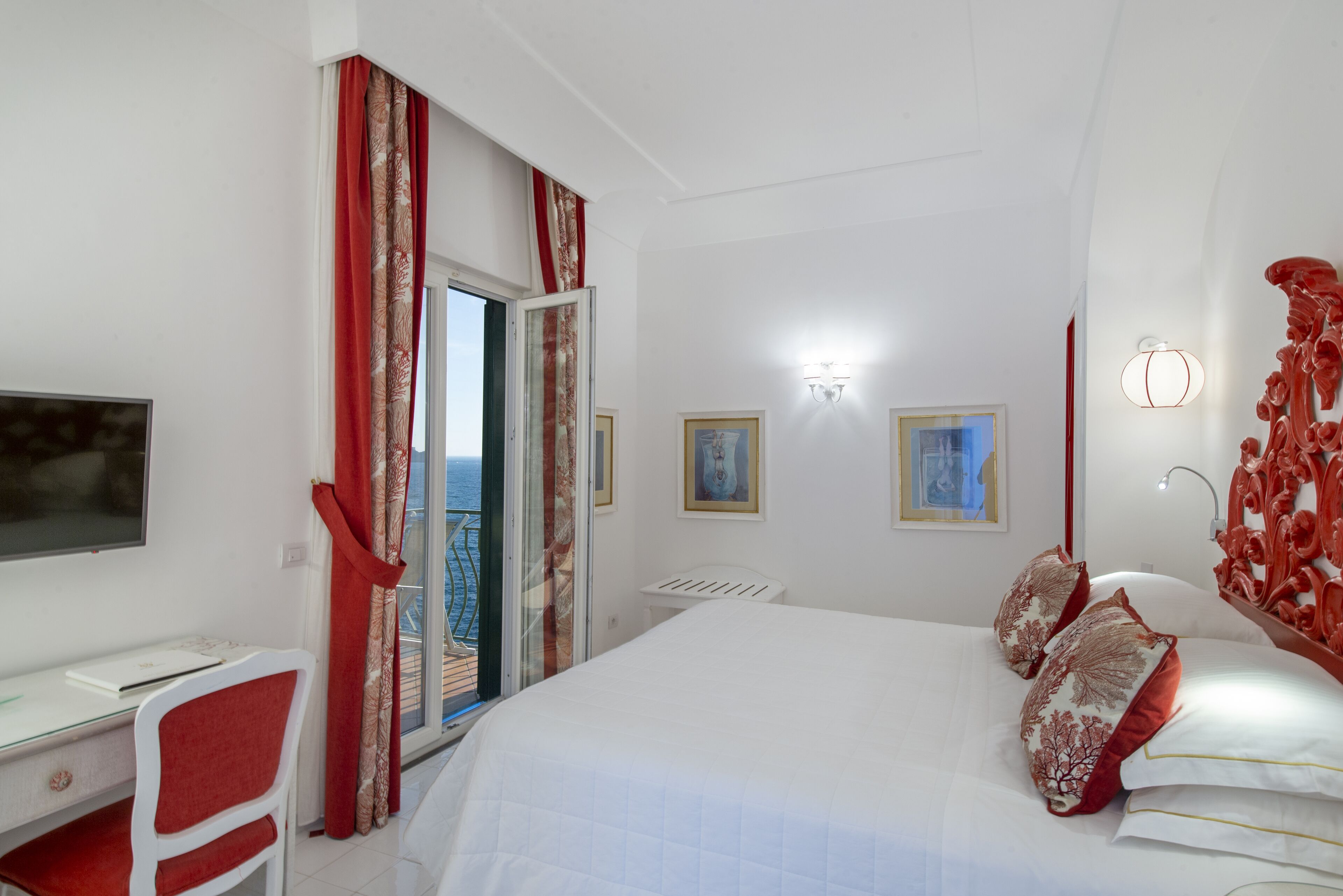 Ein Schlafzimmer mit einem Bett, einem Schreibtisch und einem Stuhl. Es gibt ein Fenster mit Vorhängen, einen an der Wand befestigten Fernseher und eine Tür, die auf einen Balkon führt.
